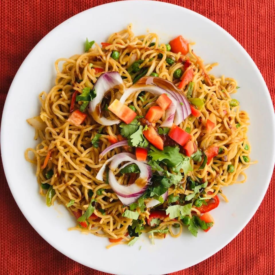 A white plate filled with noodles, red onions, bell peppers, peas, and cilantro on a red background.