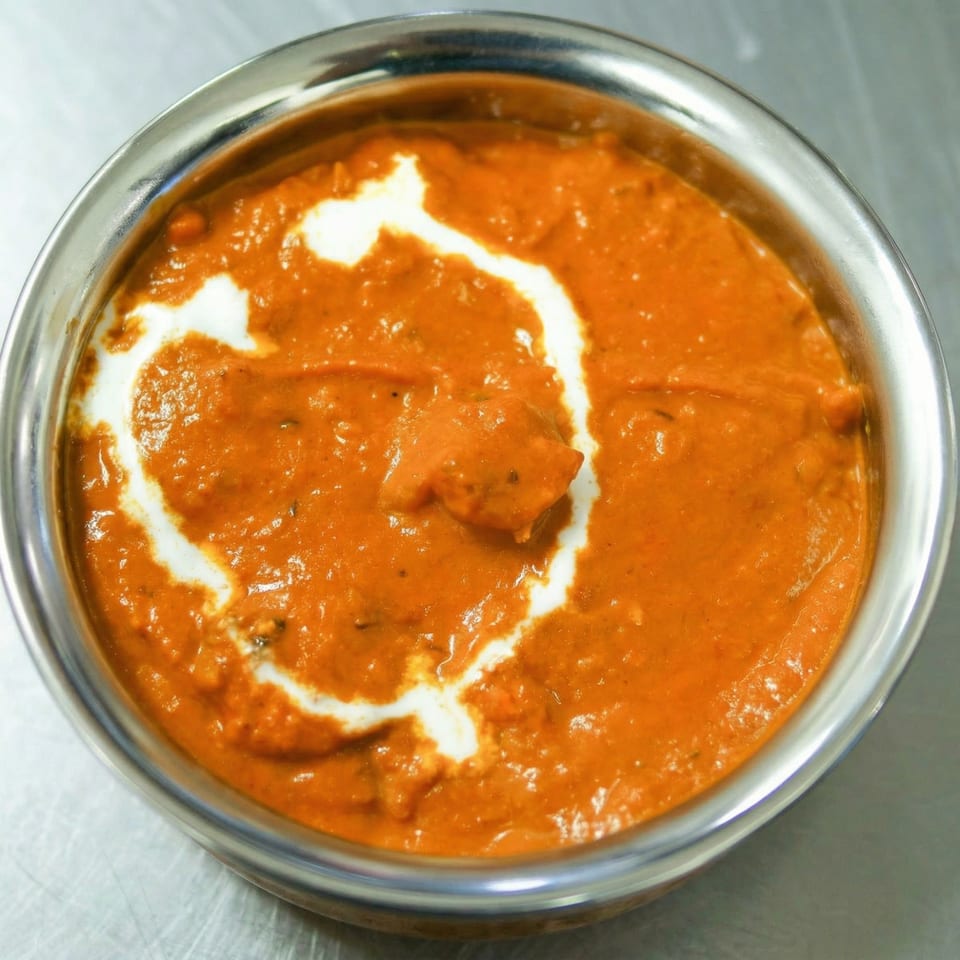Close-up of a stainless steel bowl filled with orange butter chicken, garnished with white cream swirls.