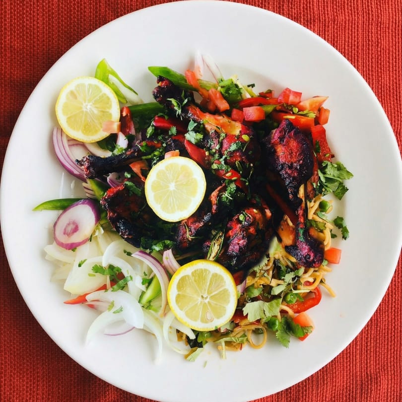 Tandoori chicken drumsticks with noodles, red onions, bell peppers, cilantro, and lemon slices.